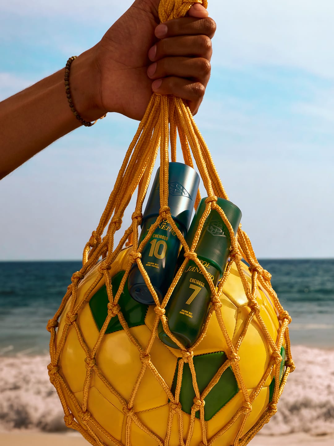 A hand holds a yellow beach ball net bag containing three green and blue sunscreen bottles with numbers 7, 8, and 10, set against a beach and ocean background.